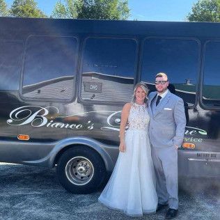 A couple in formal wedding attire stands smiling in front of a dark luxury shuttle bus labeled 