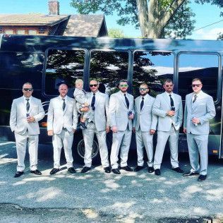 A group of eight people wearing light-gray suits and sunglasses stand in a line in front of a dark charter bus.