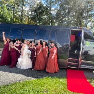 A bride and her bridesmaids in red dresses stand in front of a black party bus next to a rolled-out red carpet.