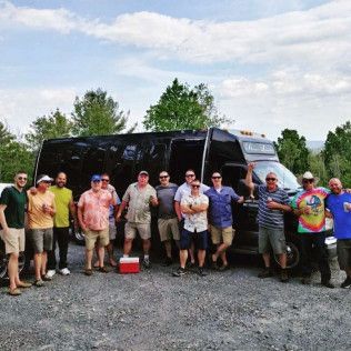 A group of people standing in front of a black passenger bus parked on a gravel lot under a bright, sunny sky.