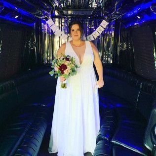 A bride in a white gown holding a bouquet inside a limo with blue accent lighting and a hanging banner.