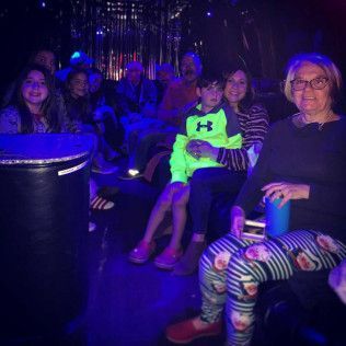 A group of people sit together inside a limousine with neon blue lighting, chatting and smiling.