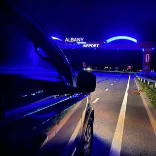 A dark night view from inside a car parked on the shoulder, showing a bright, blue-lit Albany Airport entrance sign ahead.