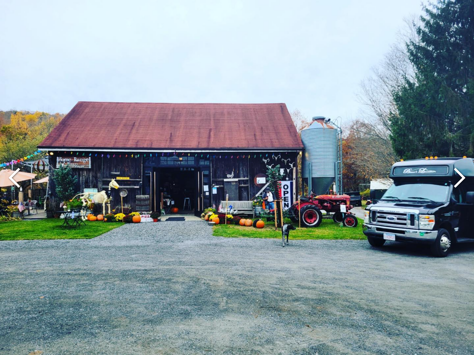 A rustic, dark-wood barn with a red metal roof, pumpkin decorations, a vintage red tractor, and a shuttle bus parked nearby.