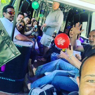 A group of people sitting inside a party bus, some holding drinks and a red birthday balloon, smiling and waving.