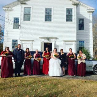 A wedding party poses for a photo in front of a white building with a long vehicle. The bridal party wears burgundy.