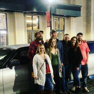 A group of seven people pose for a photo in front of a silver limousine outside a building at night.
