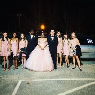 A group of eight formal wear-clad people pose for a photo at night in front of a parked limousine.