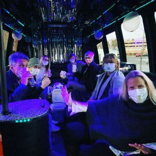 A group of people wearing face masks sit inside a dimly lit limo with blue LED lighting and metallic fringe decorations.