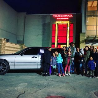 A group of people standing next to a silver limousine at night in front of a Texas de Brazil restaurant.