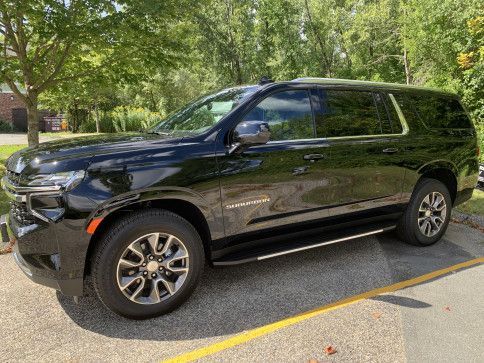 A black Chevrolet Suburban SUV parked on an asphalt lot with trees and greenery in the background.
