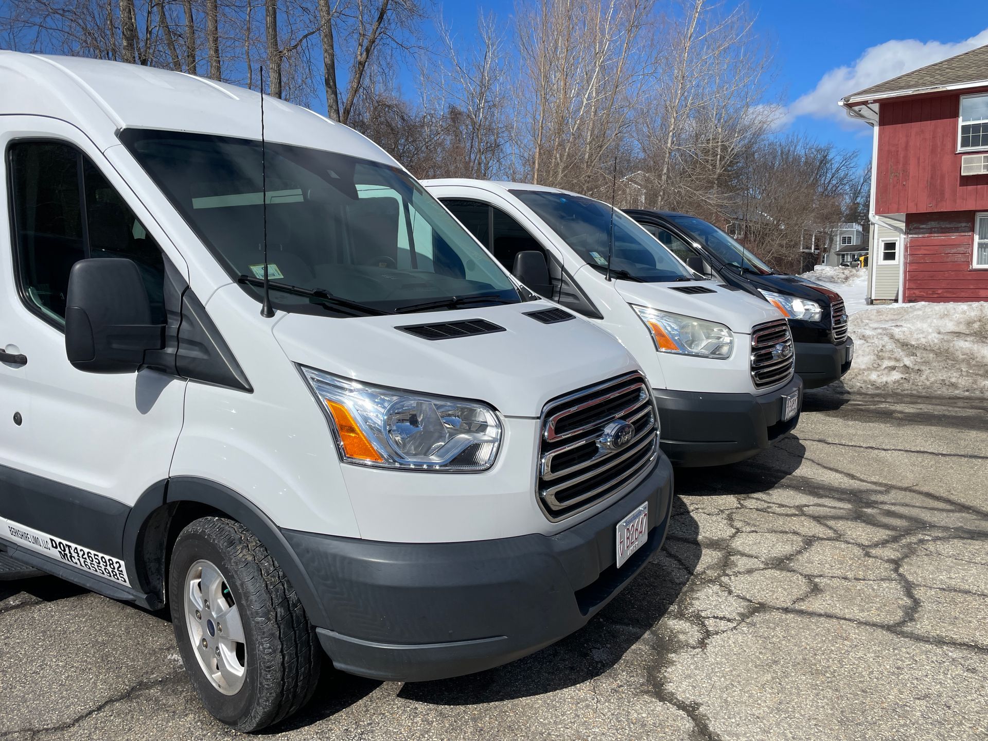 Three light-colored Ford Transit vans parked in a row on a gravel lot next to a red building on a sunny winter day.
