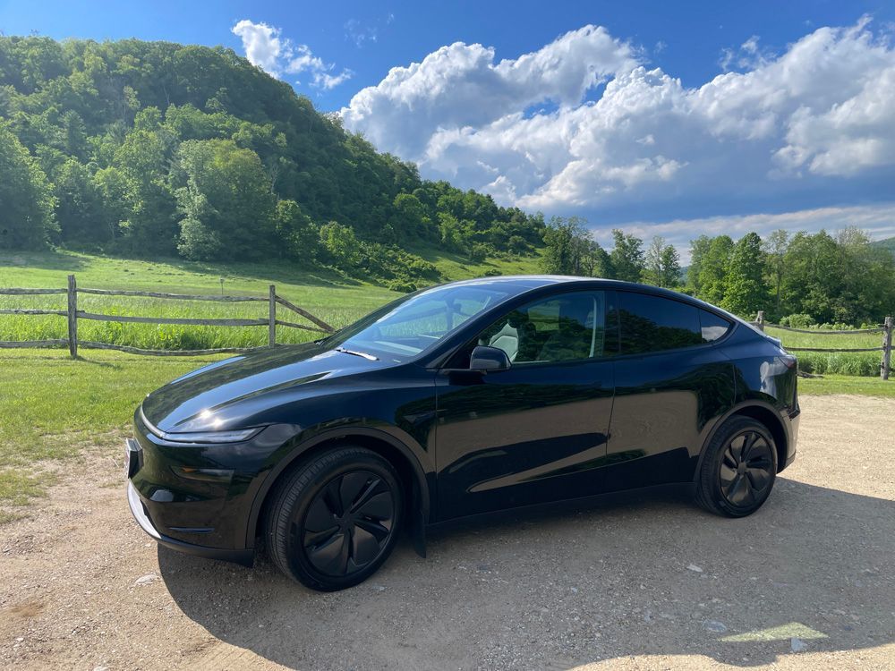 A black Tesla Model Y parked on a gravel road against a backdrop of a lush green hillside and a blue sky with clouds.