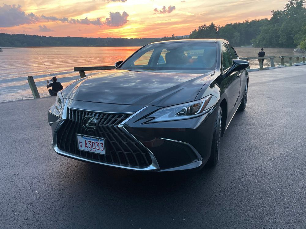 A dark Lexus sedan parked by a lake during a sunset with two people fishing in the background.