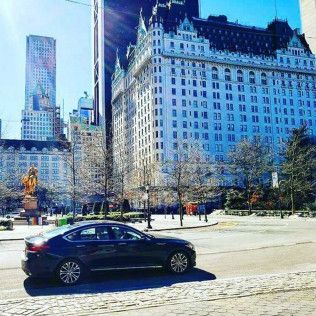A dark sedan drives on a road in front of the ornate Plaza Hotel in New York City on a bright, sunny day.