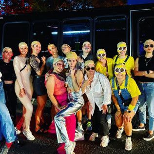 A group of people wearing yellow swim caps and creative costumes, including Minion-themed attire, pose in front of a bus.