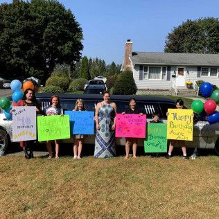 A group of people holding birthday signs stand in front of a long limousine in front of a house on a sunny day.