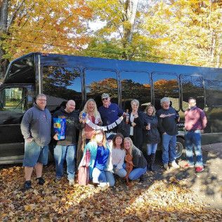 A group of people standing in front of a black shuttle bus against a background of autumn trees.