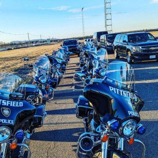 A row of parked black Pittsfield Police motorcycles, facing forward, next to a line of black SUVs on an asphalt lot.