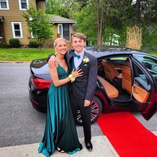 A couple in formal attire stands by a car with an open door on a red carpet in front of a residential home.