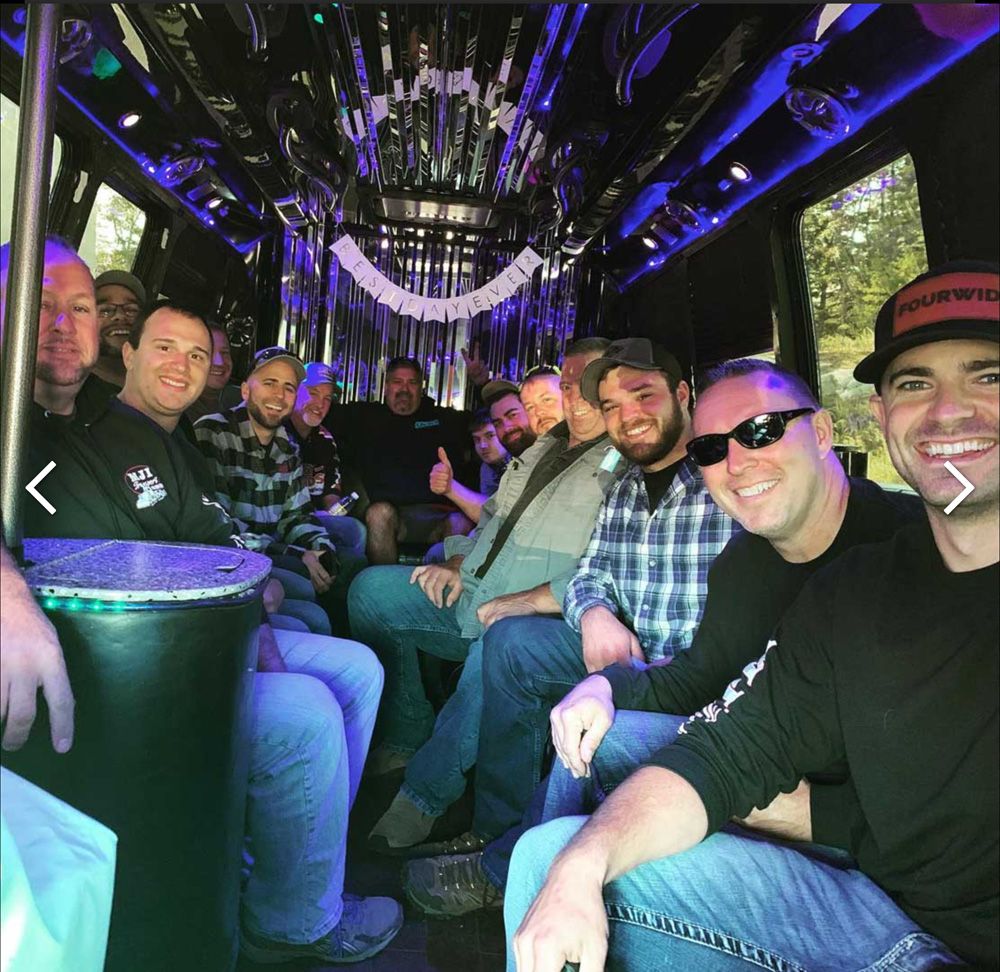 A large group of smiling friends sitting together inside a decorated party bus with purple interior lighting.