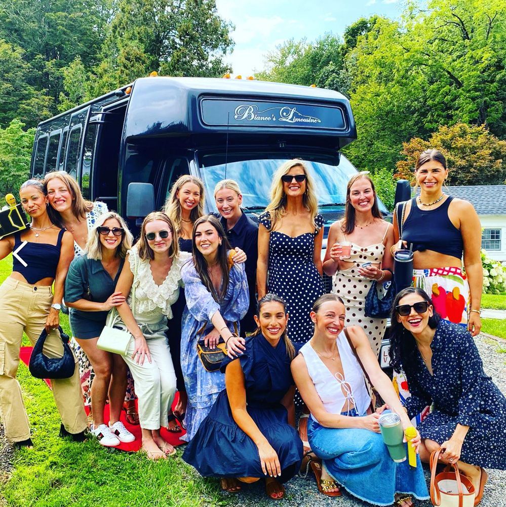 A group of 11 people posing in front of a black limousine bus on a sunny day at a grassy outdoor location.