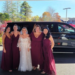 A bride in a white gown and four bridesmaids in matching dark red dresses stand smiling together by a black vehicle.