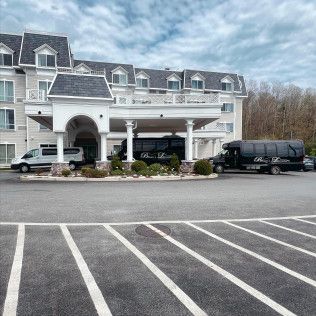 A light-colored hotel entrance with white columns, featuring a white van and a black passenger bus parked out front.