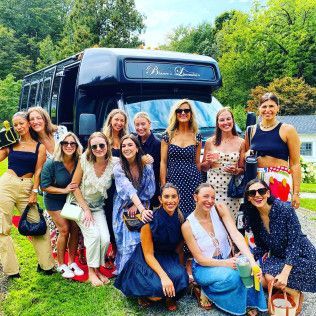 A group of women stand and pose in front of a dark bus in an outdoor, sunny setting.