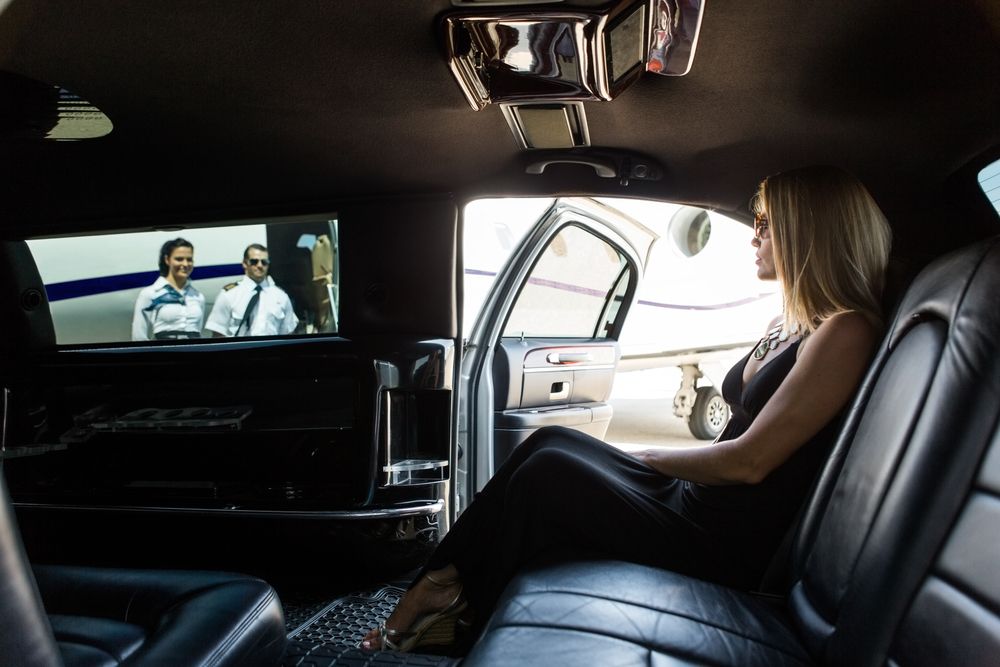 A person in a black dress sits in the back of a limousine, looking out a door at an airplane on a runway.
