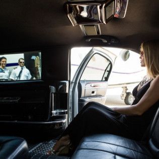 A person in a black dress sits in a limousine, looking out an open door toward an airplane on a runway.