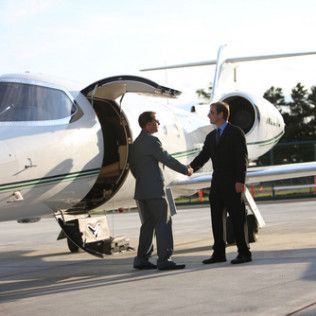 Two professionals in suits shake hands in front of a private jet on an airport tarmac.