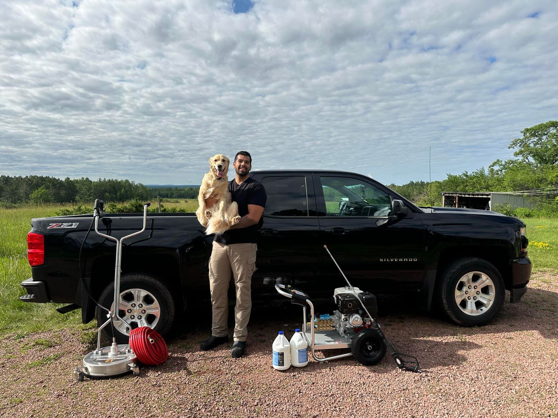 Person holding a dog beside a black pickup truck with camping gear on a gravel road under a cloudy sky
