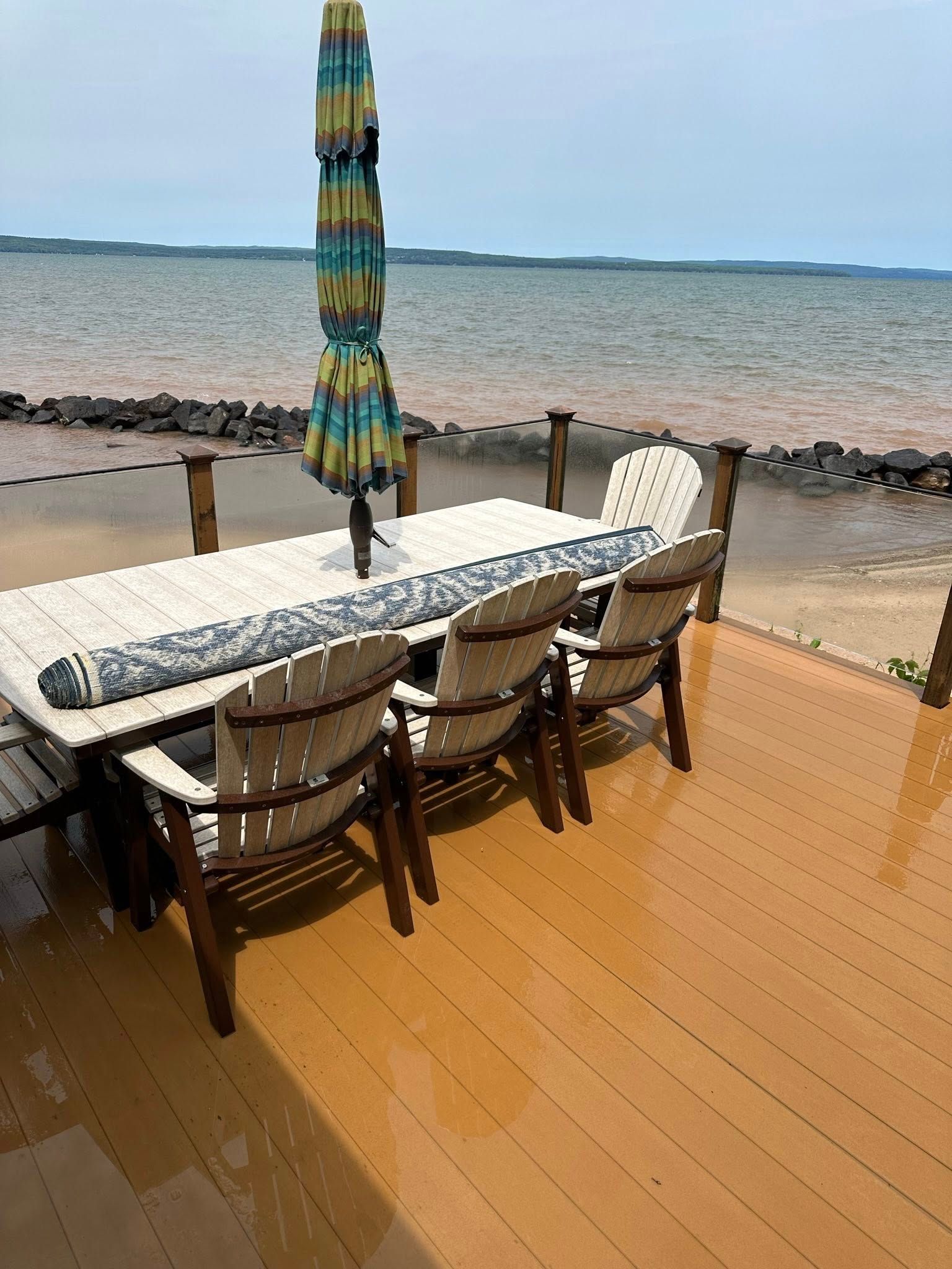 Patio table and chairs with a closed umbrella beside a rocky waterfront under a cloudy sky