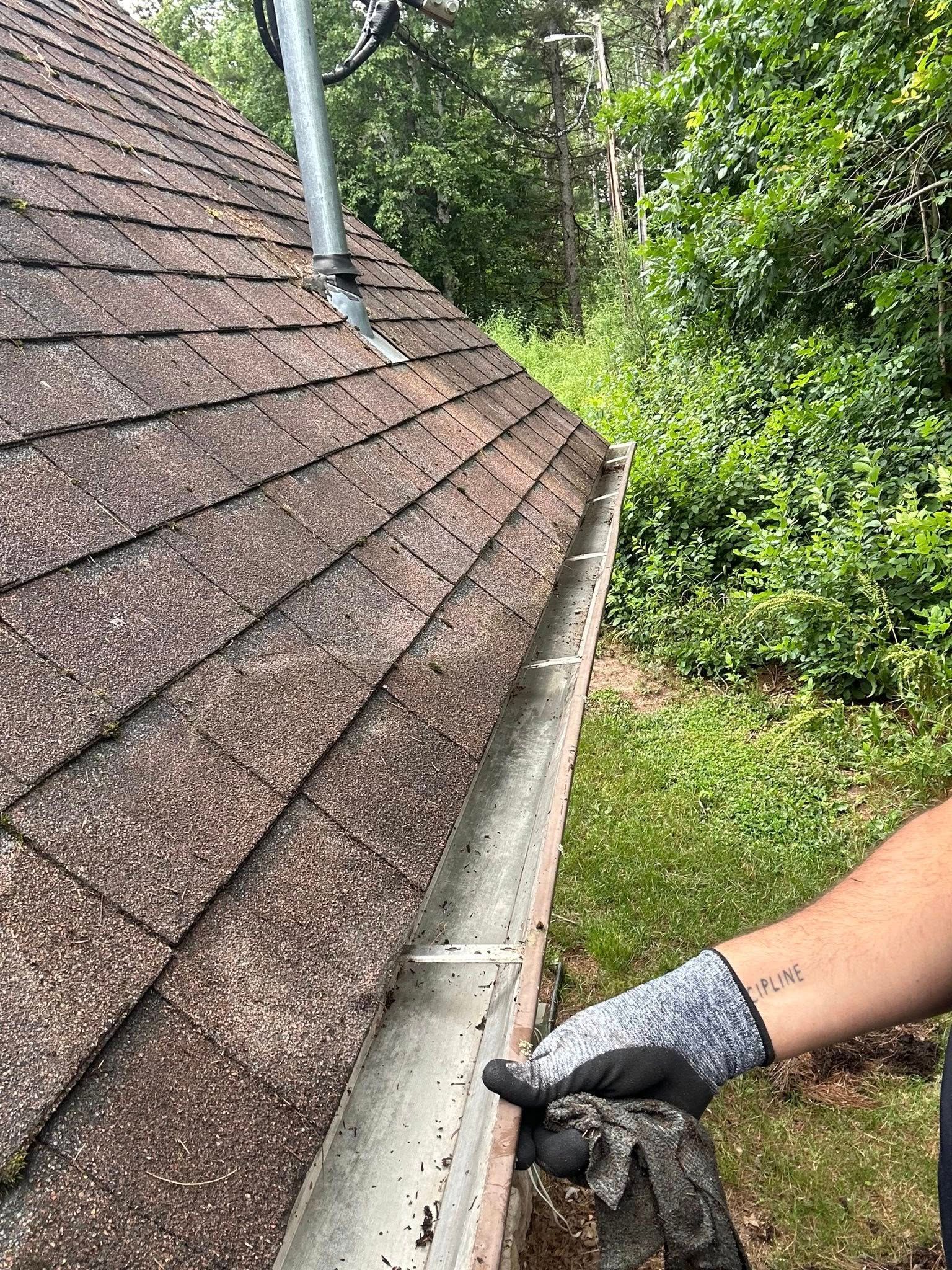 Hand with glove holding roof gutter beside a shingled roof and green yard.