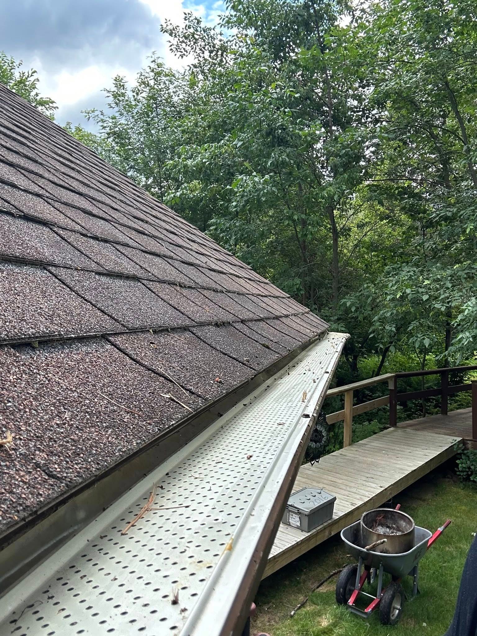 Roof gutter and downspout beside a shingled roof, with trees and a deck railing in the background