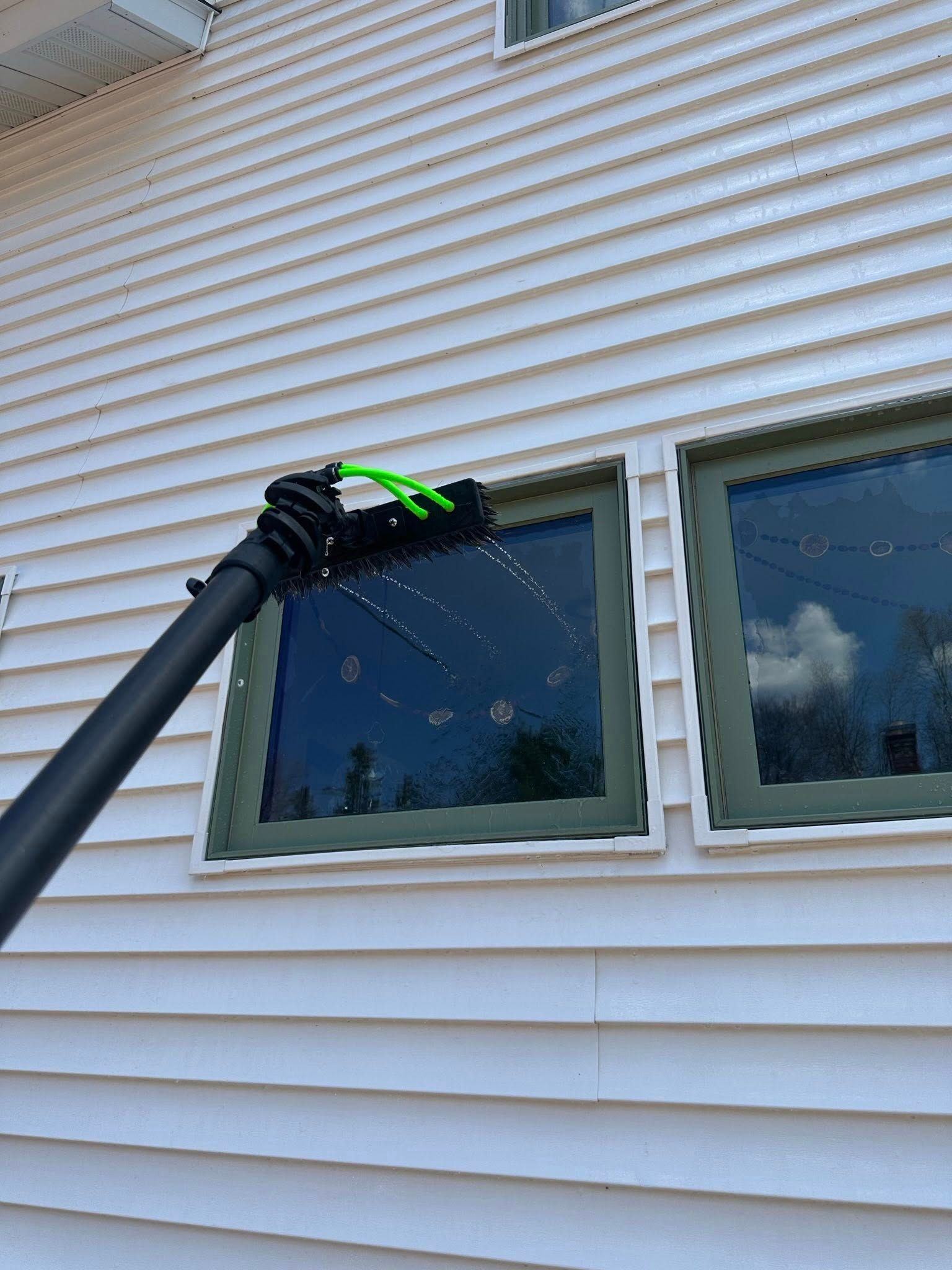 Black gloved hand on a pole cleaning a house window beneath beige siding
