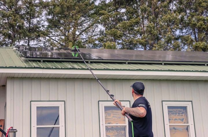 Person using a long pole to clean a green roof on a house with white siding.