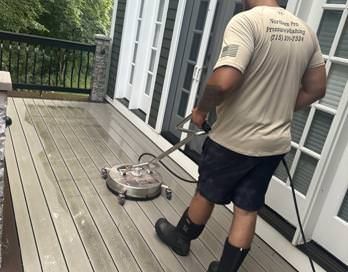 Person pressure-washing a wooden deck beside a house with a handheld cleaner