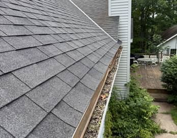 Steep gray shingle roof with a clogged gutter beside a house and walkway below