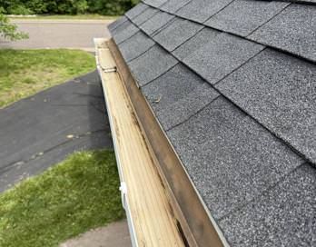 Close-up of gray shingled roof edge with exposed wooden fascia and black driveway below