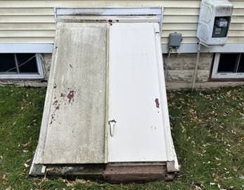 Weathered basement bulkhead doors, one white and one dirty beige, at the side of a house.