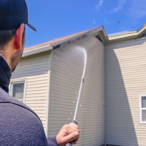 Person pressure-washing a gray house exterior under a blue sky