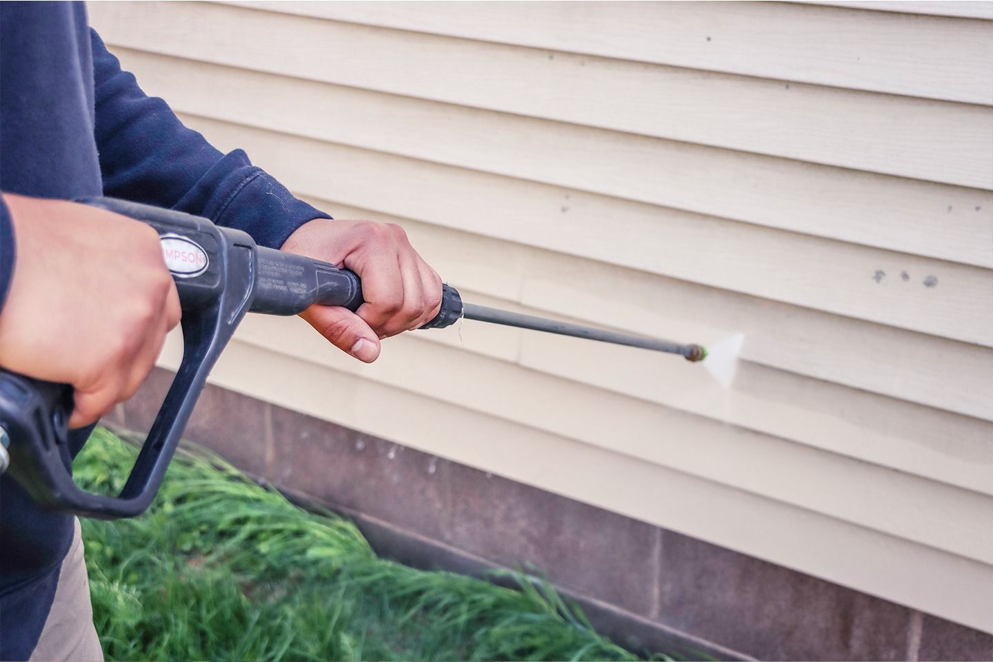 Person pressure-washing beige vinyl siding with a black spray wand.