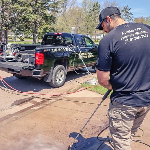 Worker using a hose to clean a black pickup truck in a driveway, with trees in the background.