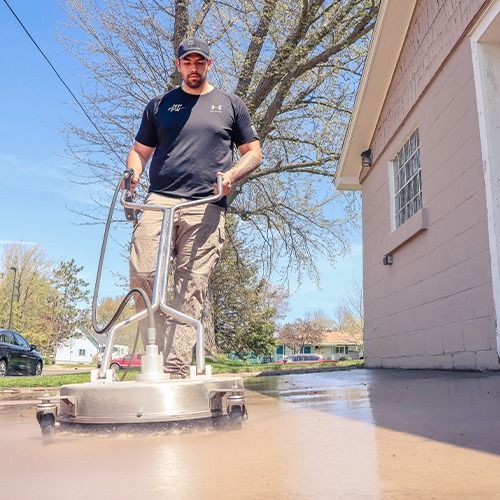 Person using a walk-behind floor scrubber on a wet driveway beside a house