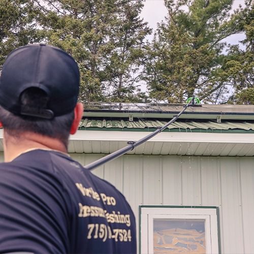 Worker pressure-washes a green roofline beside a white building with trees in the background.