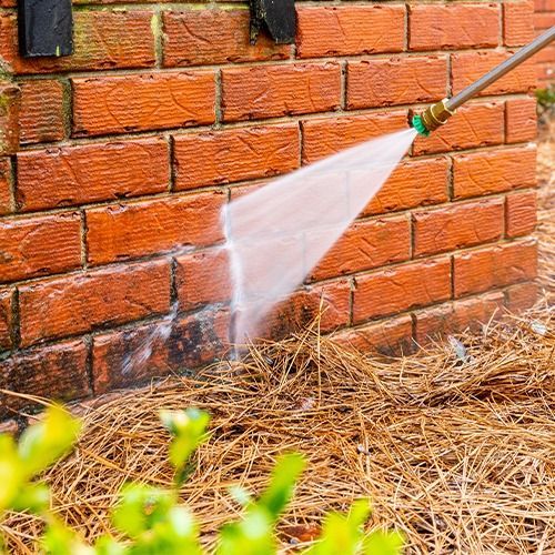 Garden hose spraying water on a red brick wall and dry mulch ground