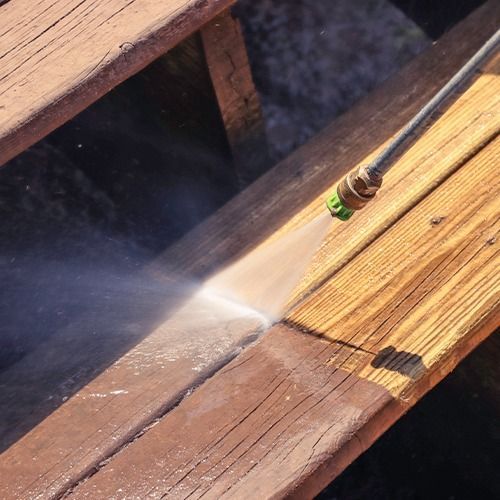 Pressure-washing a wooden deck with a hose, spraying water over sunlit boards