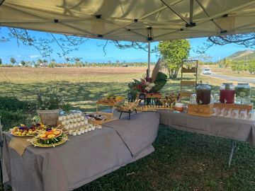 Buffet en plein air sous un auvent, avec plateaux de nourriture et de boissons, dans un champ herbeux.